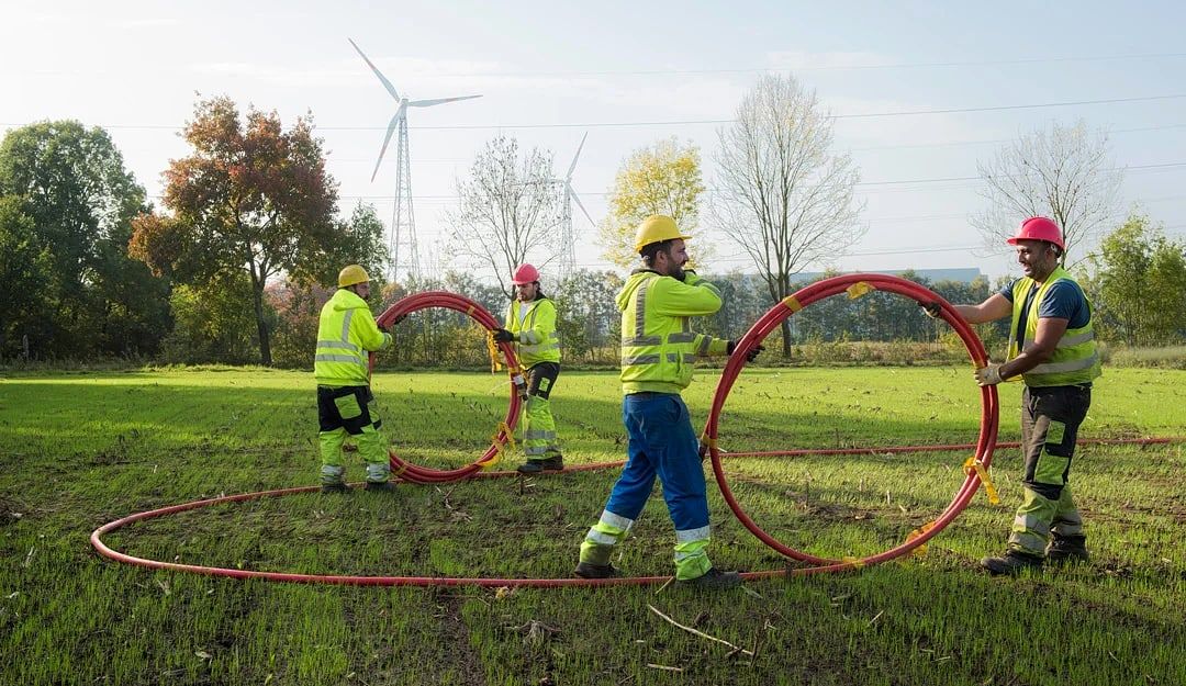 Belgisch fibre-team in hi-vis legt HDPE-buizen op een veld — windturbines en hoogspanningsmasten op de achtergrond.